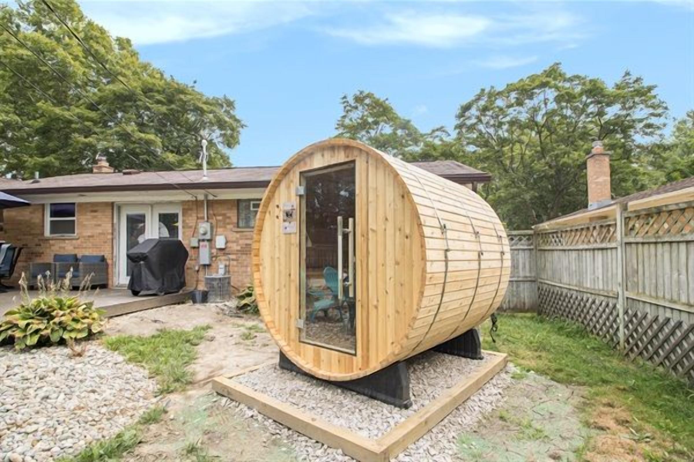 A cylindrical wooden structure with a door, possibly a sauna or hot tub, in a backyard with a house, a barbecue grill, and a wooden fence.