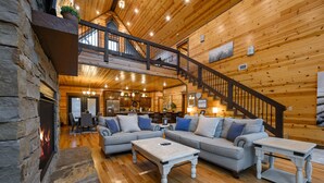 A spacious log cabin interior with a stone fireplace, a dining area, a living room with a sofa and coffee table, and a staircase leading to an upper level.