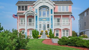 A two-story building with a central entrance, multiple balconies, and a flag on each side. - Beach Royalty - Nags Head MP 15.5, Luxury Oceanfront, Private Walk to Beach, Elevator, Pool, Hot Tub (Nags Head)