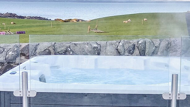 Un jacuzzi con pared de piedra de fondo, un campo de hierba con ovejas, y vista al mar y colinas en la distancia.