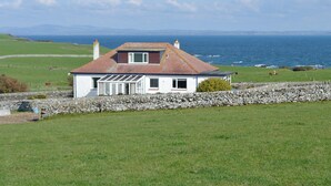 Una casa costera con un muro de piedra en primer plano, techo inclinado y vista al mar.