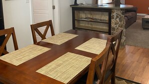 A dining area with a wooden table and chairs, a stone accent wall, a clock, and a welcome sign. - Mama Bear Cottage (Downtown Cody) (Cody)