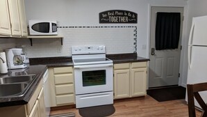 A kitchen with white cabinets, a microwave, a stove, and a refrigerator. There is a sign on the wall that says "The Best Place To Be Is Together". - Mama Bear Cottage (Downtown Cody) (Cody)