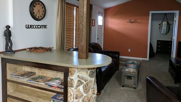 A living room with a stone pillar, a clock, a 'Welcome' sign, a ceiling fan, and a brown leather couch. - Mama Bear Cottage (Downtown Cody) (Cody)