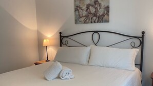 A neatly made bed with white linens, a painting of horses on the wall, and a lamp beside the bed. - Mama Bear Cottage (Downtown Cody) (Cody)