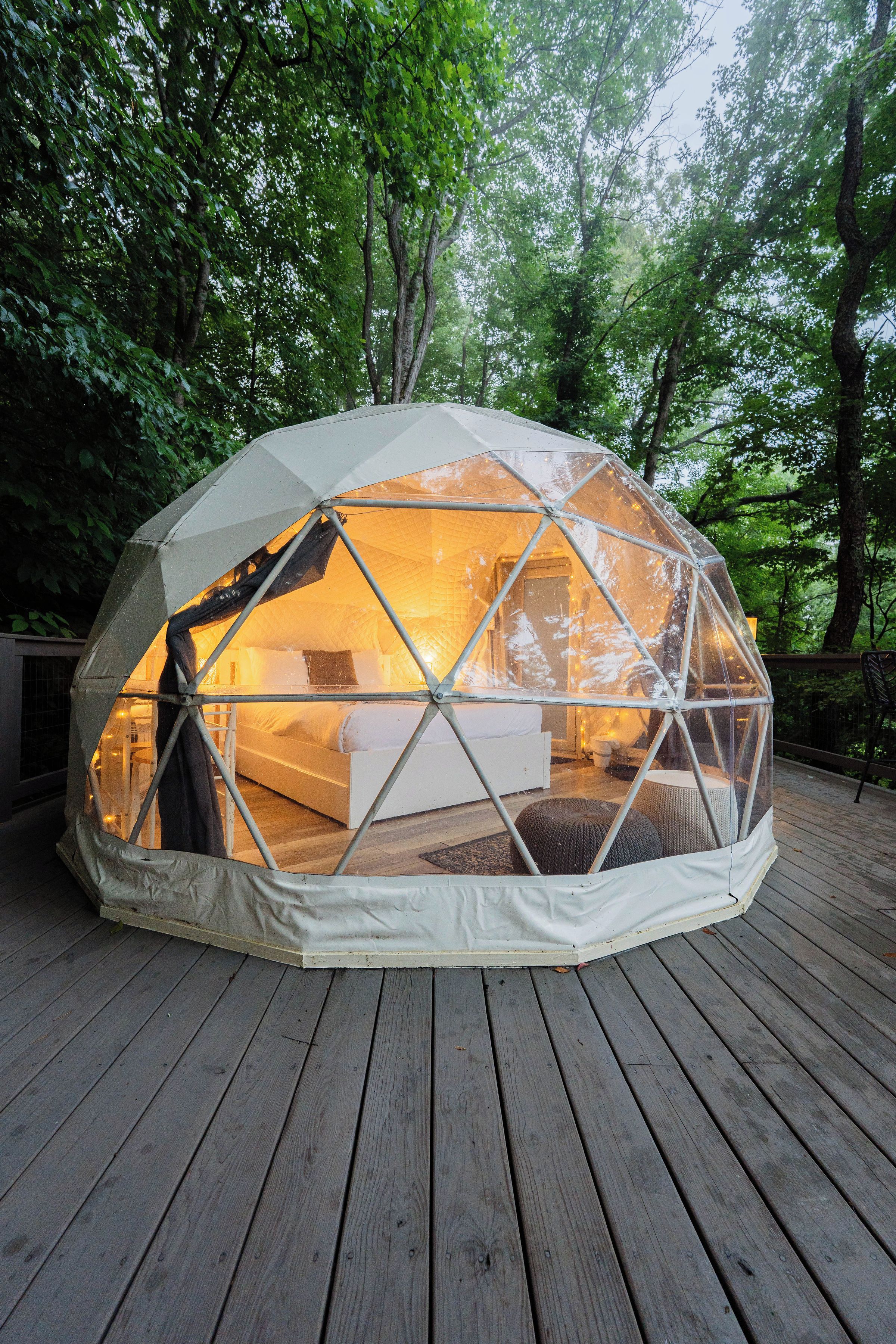 Eine geodätische Kuppel mit einem Bett im Inneren, umgeben von einer Holzterrasse und Bäumen.