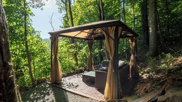 Um gazebo de madeira com um banco e uma mesa, rodeado por árvores.