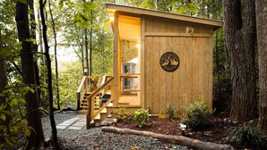 A wooden cabin with a glass door, surrounded by trees and gravel. - Hot Tub, Views, Outdoor Oasis (Todd)