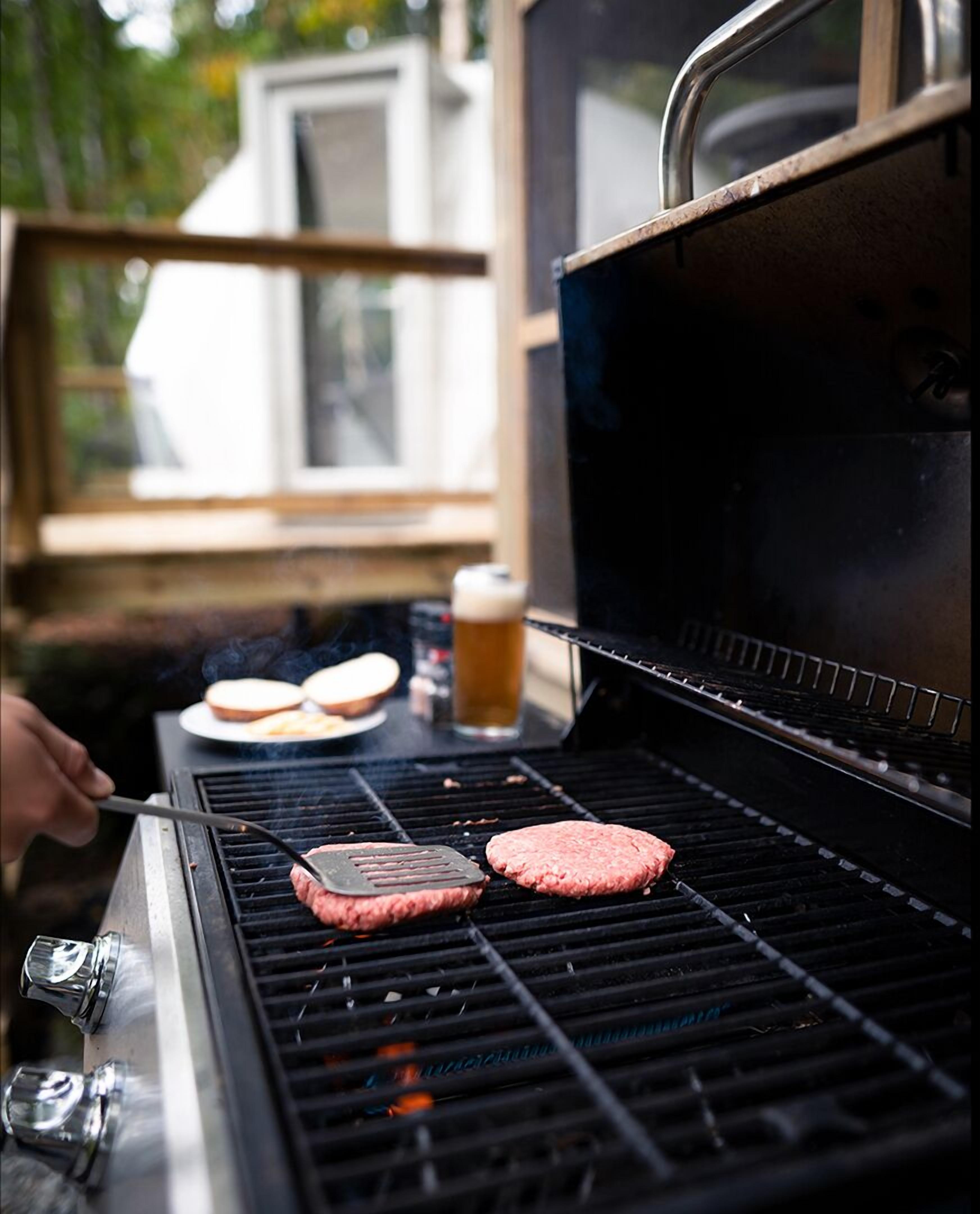 A grill with two hamburgers cooking, a beer, and a sandwich on a table.