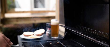 A grill with two hamburgers cooking, a beer, and a sandwich on a table.