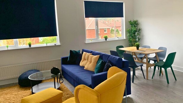 A modern living room with a blue sofa, yellow armchair, and a dining table with chairs. There are two windows with blinds, a radiator, and a small plant on the windowsill. - Consett Shotley Bridge, Stylish & Modern Two Bedroom Apartment (Consett)