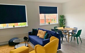 Un salon moderne avec un canapé bleu, un fauteuil jaune et une table à manger avec des chaises. Il y a deux fenêtres équipées de stores, un radiateur et une petite plante posée sur le rebord de la fenêtre.