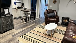 A modern living room with a leather sofa, a dining area with a table and stools, a fireplace, and a kitchen with a TV and coffee maker. - The Cedar Suite (Yellowhead County)