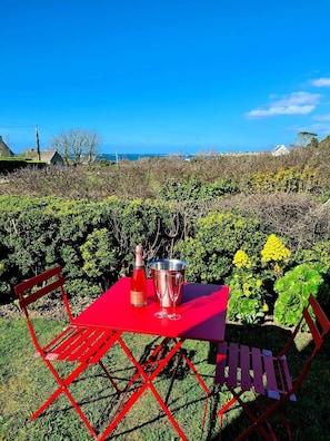 Une table rouge avec une bouteille et deux verres, entourée de verdure et offrant une vue sur la mer.