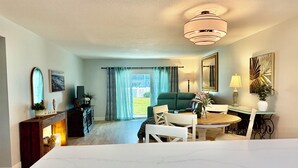 A modern living room with a green sofa, a dining table with chairs, a fireplace, and a large window with curtains. - 	Modern luxury meets coastal comfort-1200 sq.ft  completely remodeled. Pool (Ormond Beach)