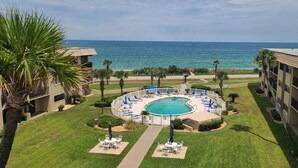 A circular pool with lounge chairs and umbrellas, surrounded by a well-maintained lawn and palm trees, with a view of the ocean. - 	Modern luxury meets coastal comfort-1200 sq.ft  completely remodeled. Pool (Ormond Beach)