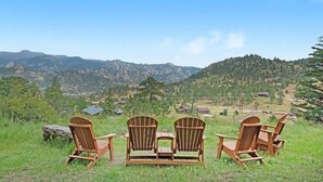 Trois fauteuils Adirondack en bois et une petite table sont placés sur une colline herbeuse, d’où l’on a une vue sur une vallée bordée de montagnes et parsemée de maisons.