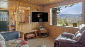 L’intérieur d’un chalet en bois est aménagé avec un téléviseur, un coin salon et une grande fenêtre donnant sur un paysage montagneux.