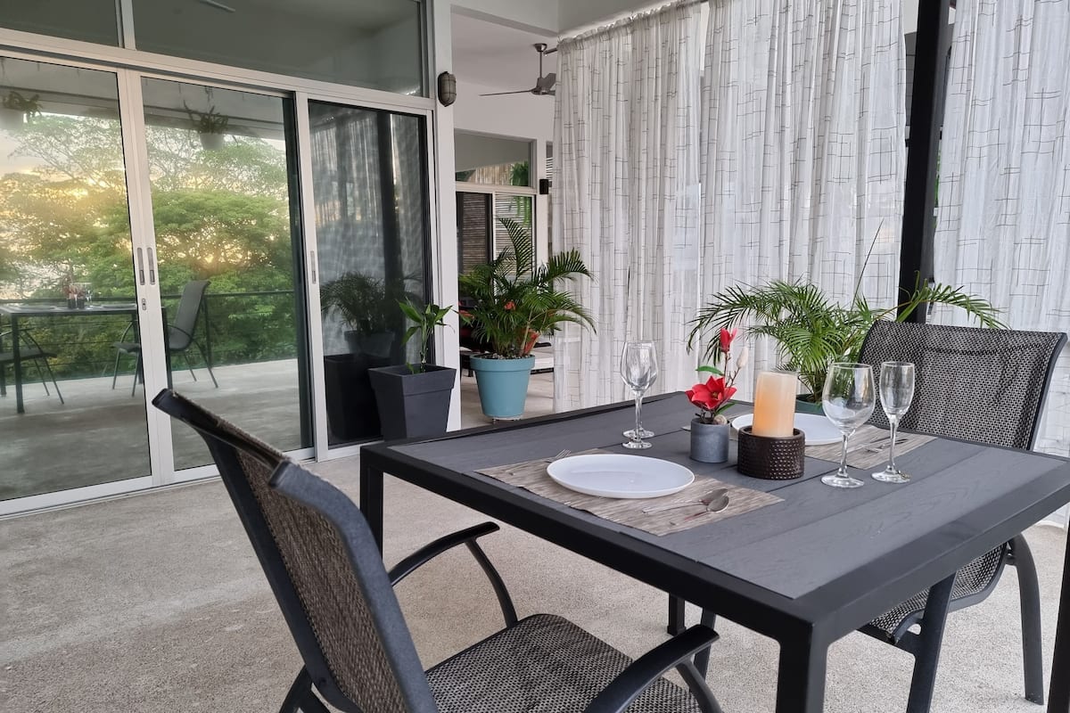 A dining table set for two with chairs, a candle, and a glass on a balcony with potted plants and a view of greenery.