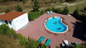 Una piscina al aire libre con borde curvo, rodeada por una zona pavimentada con sillones de descanso y un pequeño edificio de techo rojo.