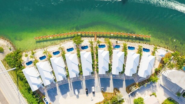 An aerial view of a resort with multiple overwater bungalows arranged in a semi-circle, connected by a wooden walkway, with a boat visible on the water.