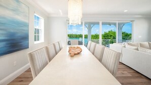 A spacious dining area with a long table, white upholstered chairs, a chandelier, and a view of a pool and greenery through large windows.