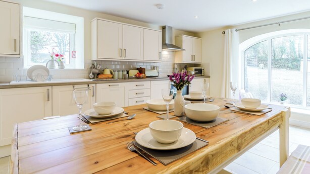 A modern kitchen with white cabinets, a wooden dining table set for four, and a view of the garden through large windows.