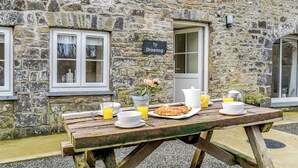 A stone building with a sign reading 'Tŷ Drenog,' a wooden table with breakfast items, and a window with a view.