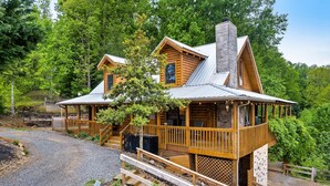 A log cabin with a porch, stone chimney, and a view of the surrounding forest. - Drive in Theater! Putt-Putt | Fire Pit | Top 10% (Sevierville)