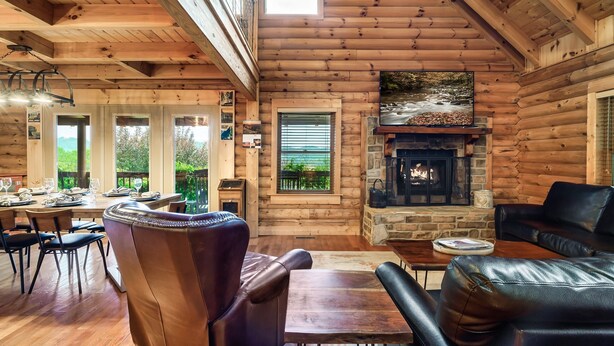 A cozy log cabin interior with a dining area, leather seating, a fireplace, and large windows offering views of the outdoors. - Drive in Theater! Putt-Putt | Fire Pit | Top 10% (Sevierville)