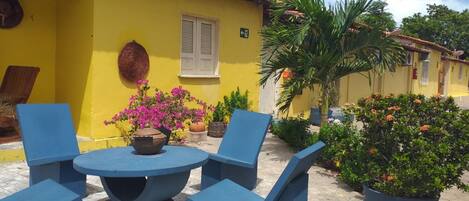 A yellow building with a tiled roof, a round table with blue chairs, and potted plants.