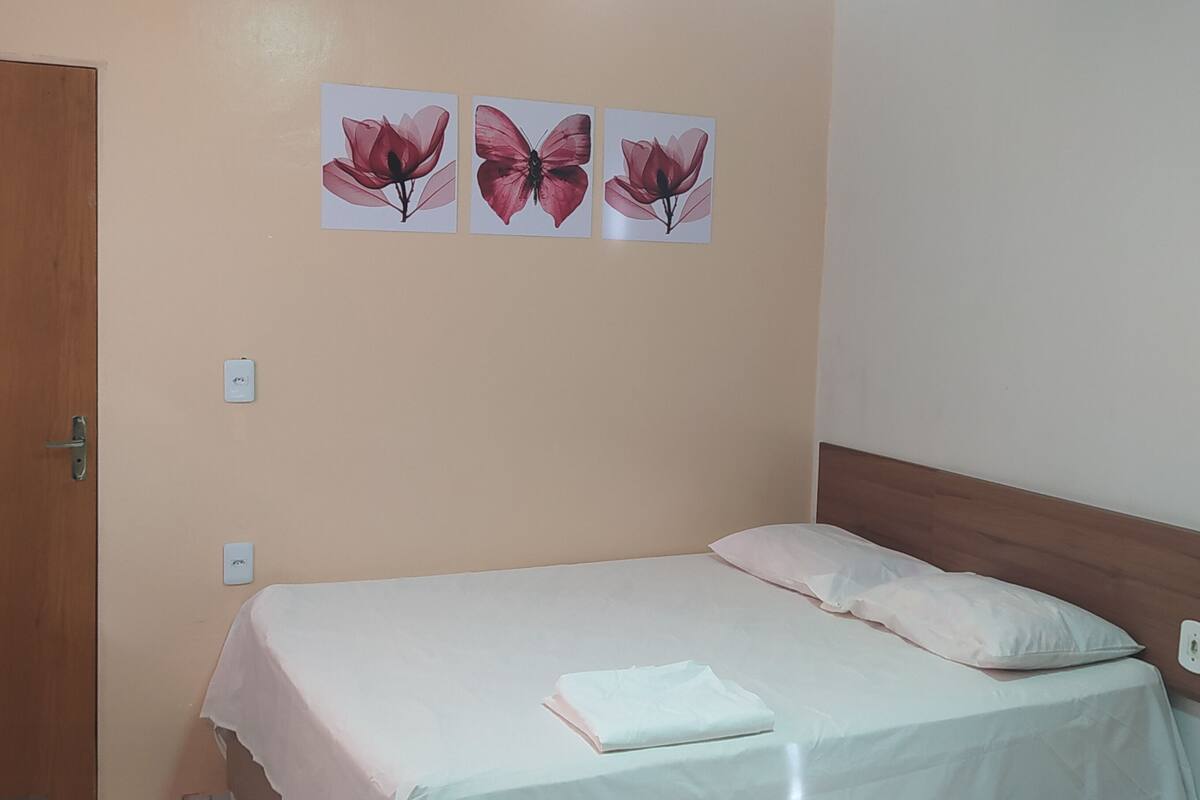 A single bed with a wooden headboard, a white pillow, and a folded white towel. There are three wall-mounted light switches and two wall-mounted hooks.