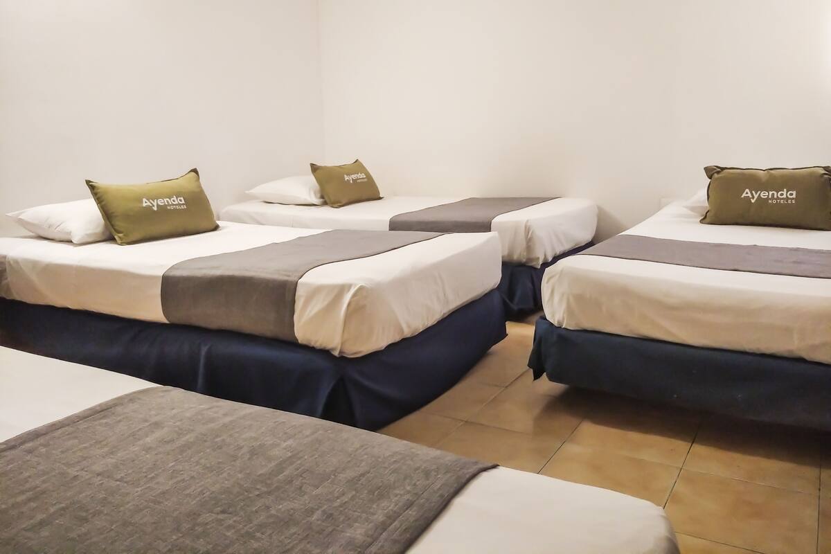 Een hotelkamer met drie enkele bedden, elk voorzien van een kussen met de tekst 'Ayenda Hotels'. De bedden hebben wit en grijs beddengoed en de kamer heeft een betegelde vloer.
