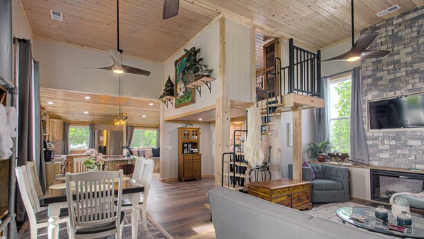 A spacious living area with a high ceiling, wooden beams, a fireplace, a TV mounted on a stone wall, and a dining area with a wooden table and chairs.