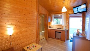 Interior de una cabaña de madera con kitchenette, una mesita con un cuadro y un televisor montado en la pared.