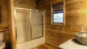 A wooden cabin bathroom with a glass shower enclosure, a toilet, and a window providing a view of trees. - LUXURY LOG CABIN #1 ON THE FRIO RIVER (Leakey)