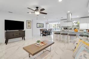 Una sala moderna con un televisor de pantalla plana, un comedor con mesa y sillas, una cocina con barra y un ventilador de techo.