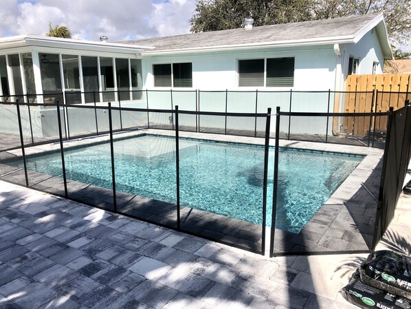 A rectangular swimming pool with a black safety fence, surrounded by a paved deck.