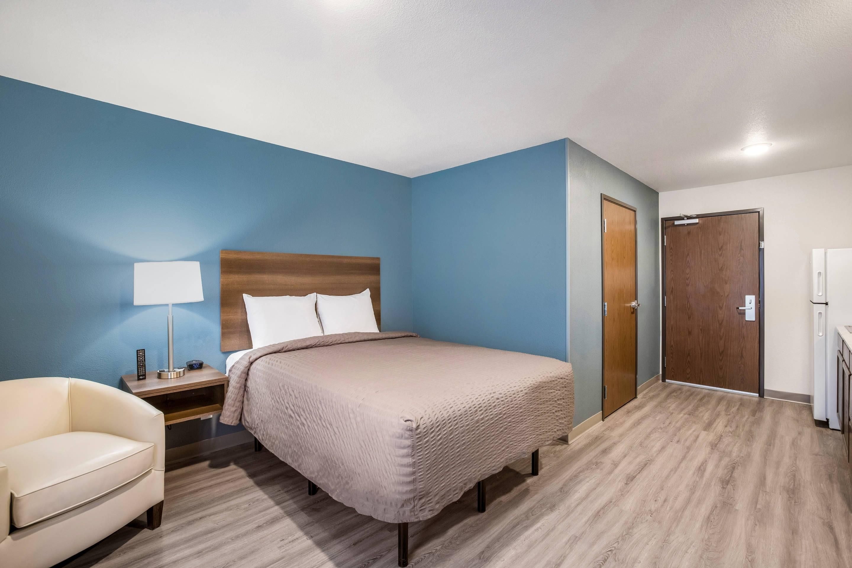 A hotel room with a bed, a chair, a nightstand with a lamp, a wooden door, and a white refrigerator.
