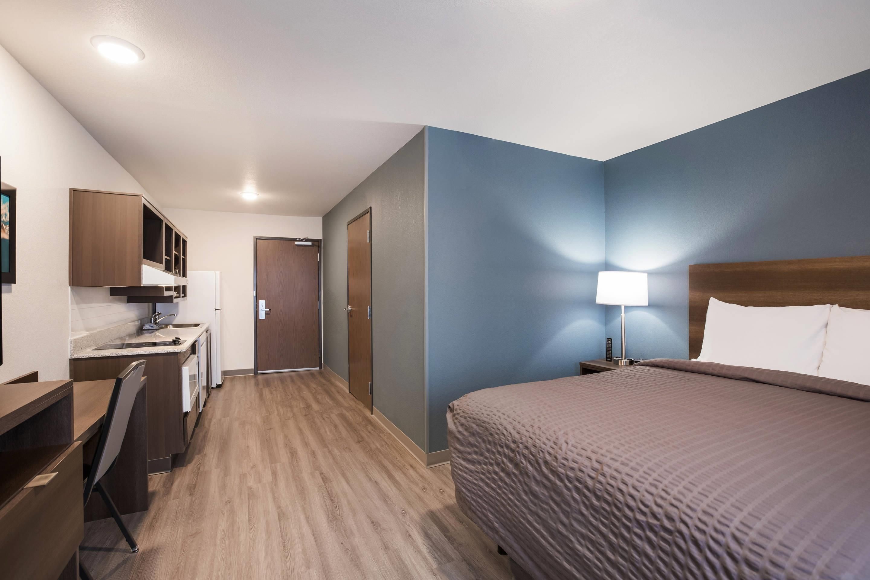 A hotel room with a bed, desk, kitchenette, and a door leading to another room.