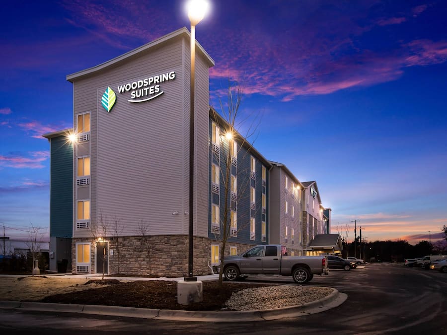 A hotel building with a sign reading 'WOODSPRING SUITES' and a parking lot with vehicles.