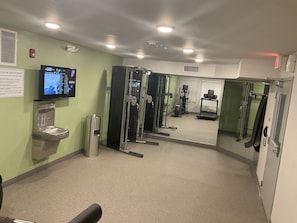 A fitness center with exercise equipment, a water dispenser, and a television displaying safety rules. - WoodSpring Suites Detroit Madison Heights (Madison Heights)