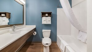 A modern bathroom with a white sink, toilet, and bathtub. There are wooden shelves with towels and a shower curtain. - WoodSpring Suites Charlotte - University Research Park (Charlotte)