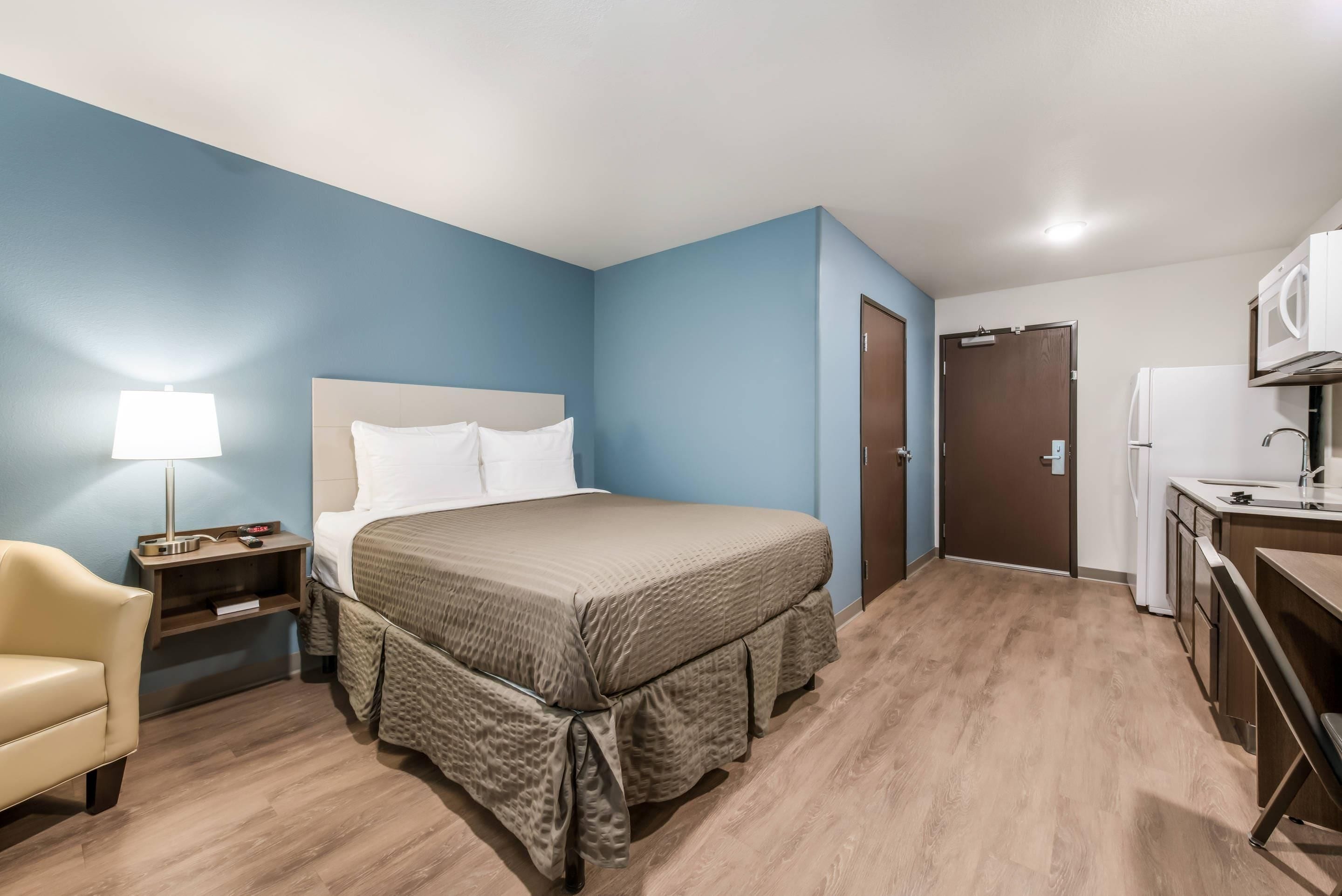 Ein Hotelzimmer mit Bett, Nachttisch, Lampe, Stuhl und einer Küchenzeile.
