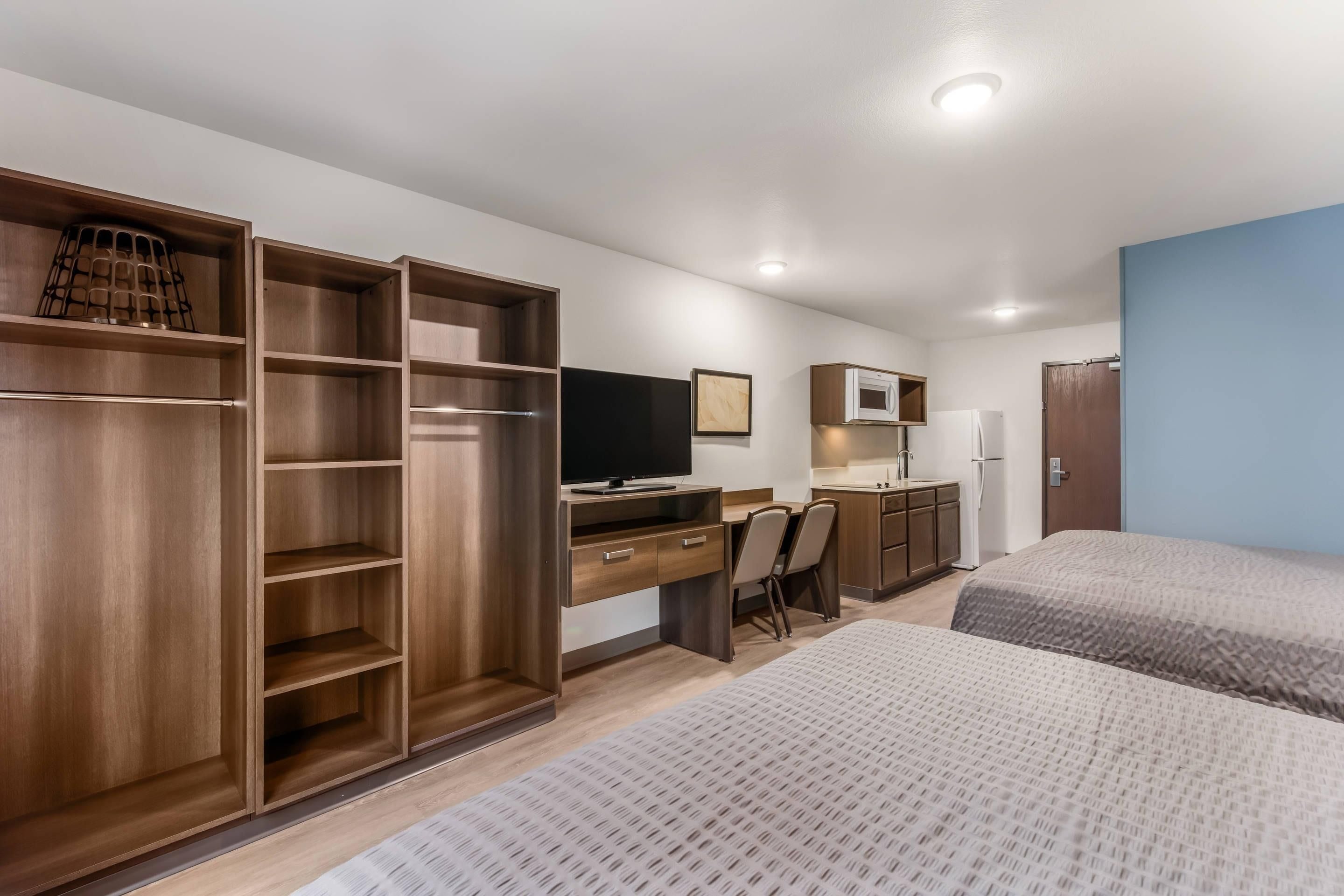 A hotel room with a wooden wardrobe, a desk with a chair, a bed, a microwave, and a refrigerator.