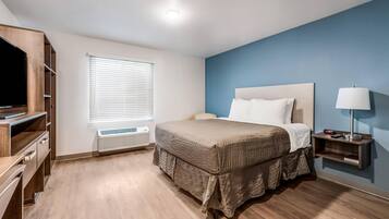 A hotel room with a bed, a TV, a heater, a window with blinds, and a bedside table with a lamp.