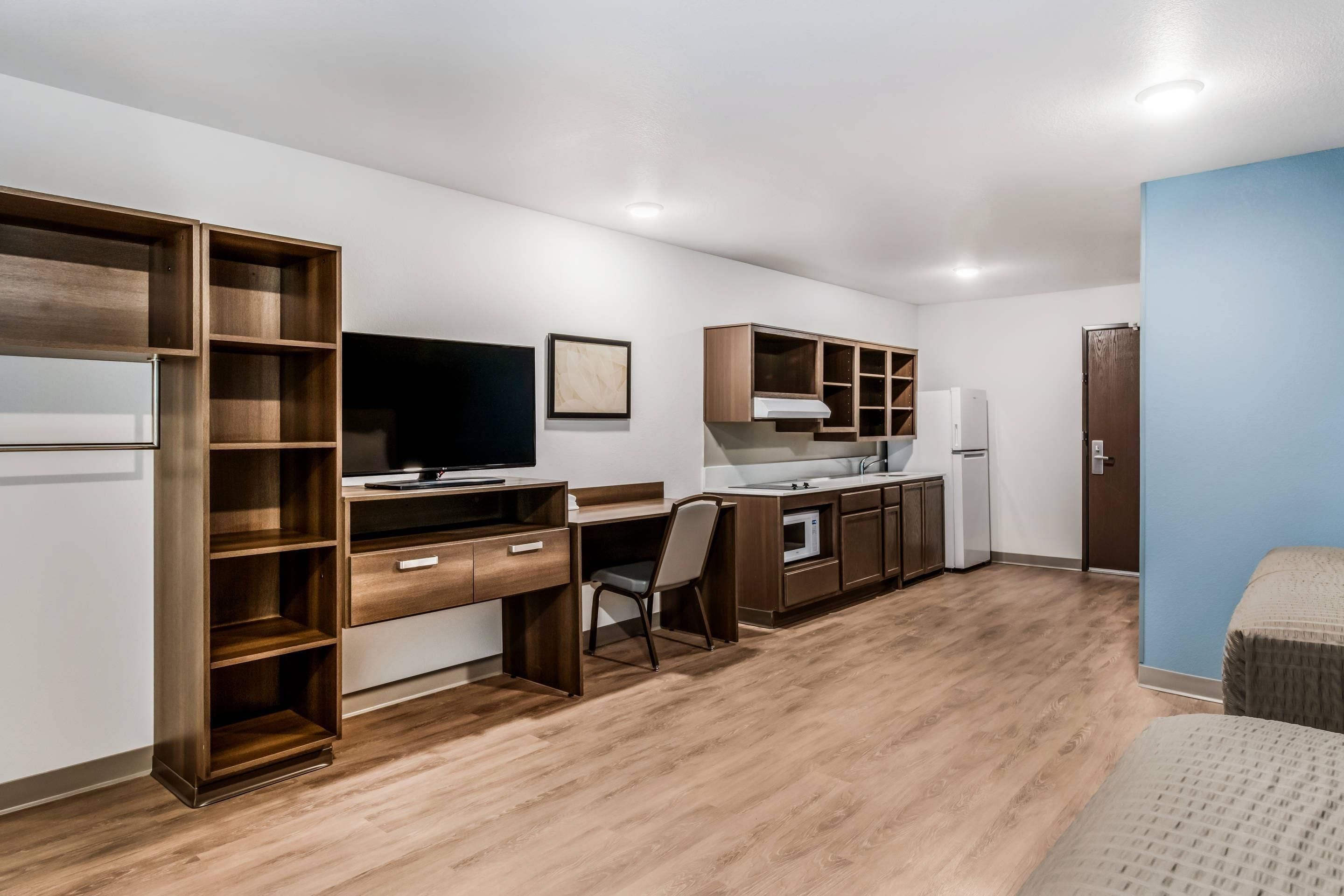 Une chambre de taille modeste, équipée d’un bureau en bois, d’une télévision, d’une kitchenette et d’une armoire.