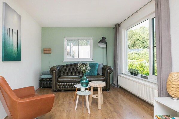 Un salon avec un canapé vert, un fauteuil en cuir marron, une petite table sur laquelle est posé un vase, et une fenêtre avec des rideaux.