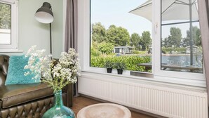 A room with a large window, a leather sofa with a blue throw pillow, a green vase with flowers, and a wooden stool. - Picturesque holiday home with garden on open waters (Terherne)