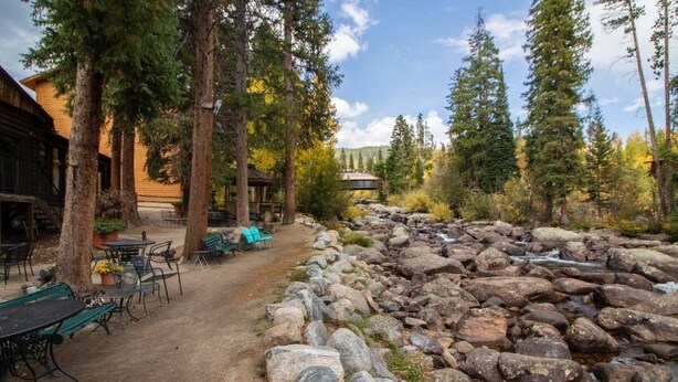 A rustic outdoor seating area with tables and chairs, a wooden building, and a rocky stream. - Riverfront condo #3 w/patio, 1block 2 town,King bed,Wifi,20 min ski Granby Ranch (GRAND LAKE)